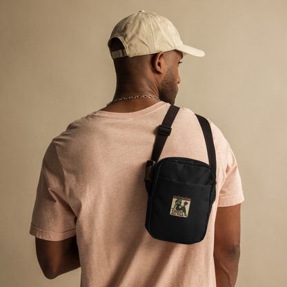 Man wearing a beige cap and light pink shirt with a black crossbody bag on a beige background. The design features a green pinup-style zombie girl holding flowers, with a banner underneath that reads “CREEPY ALL YEAR.”