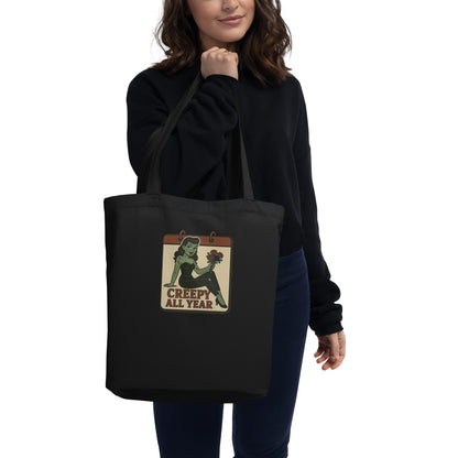 Female with brown hair holding a black tote with a vintage-style design on the pocket area. The design features a green pinup-style zombie girl holding flowers, with a banner underneath that reads “CREEPY ALL YEAR.”