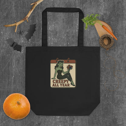 Black tote with a vintage-style patch on the front area laying on a grey background with a pumpkin and bats. The design features a green pinup-style zombie girl holding flowers, with a banner underneath that reads “CREEPY ALL YEAR.”