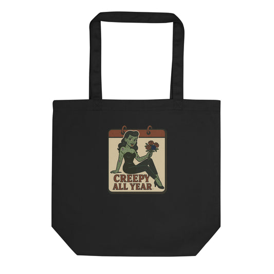 A black tote with a vintage-style design. The design features a green pinup-style zombie girl holding flowers, with a banner underneath that reads “CREEPY ALL YEAR.”