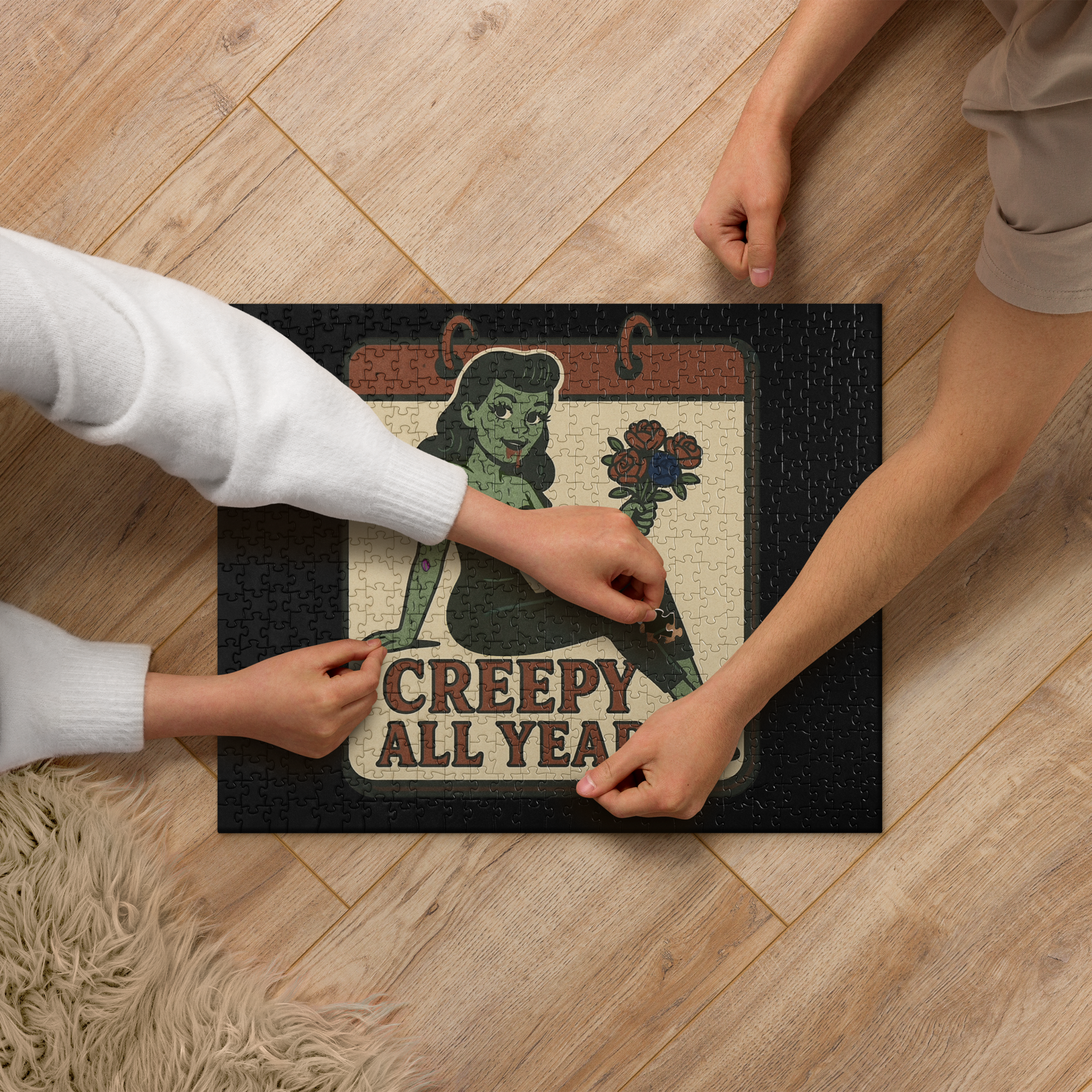 Person interacting with a puzzle featuring design on a wooden floor. The design features a green pinup-style zombie girl holding flowers, with a banner underneath that reads “CREEPY ALL YEAR.”