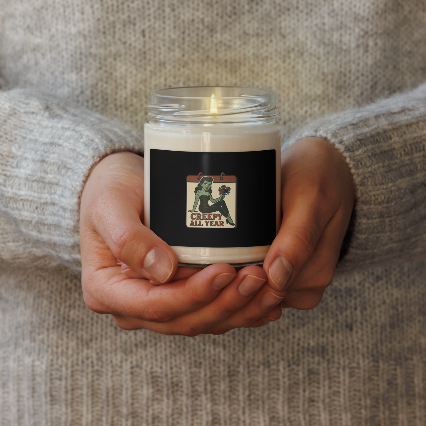 Candle with a label held in hands against a sweater and the label is a pinup zombie holding flowers and text reads Creepy All Year