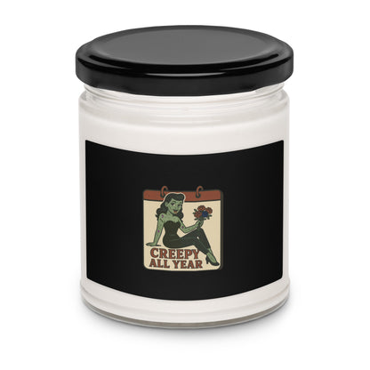 White glass candle with black metal lid. The design features a green pinup-style zombie girl holding flowers, with a banner underneath that reads “CREEPY ALL YEAR.”