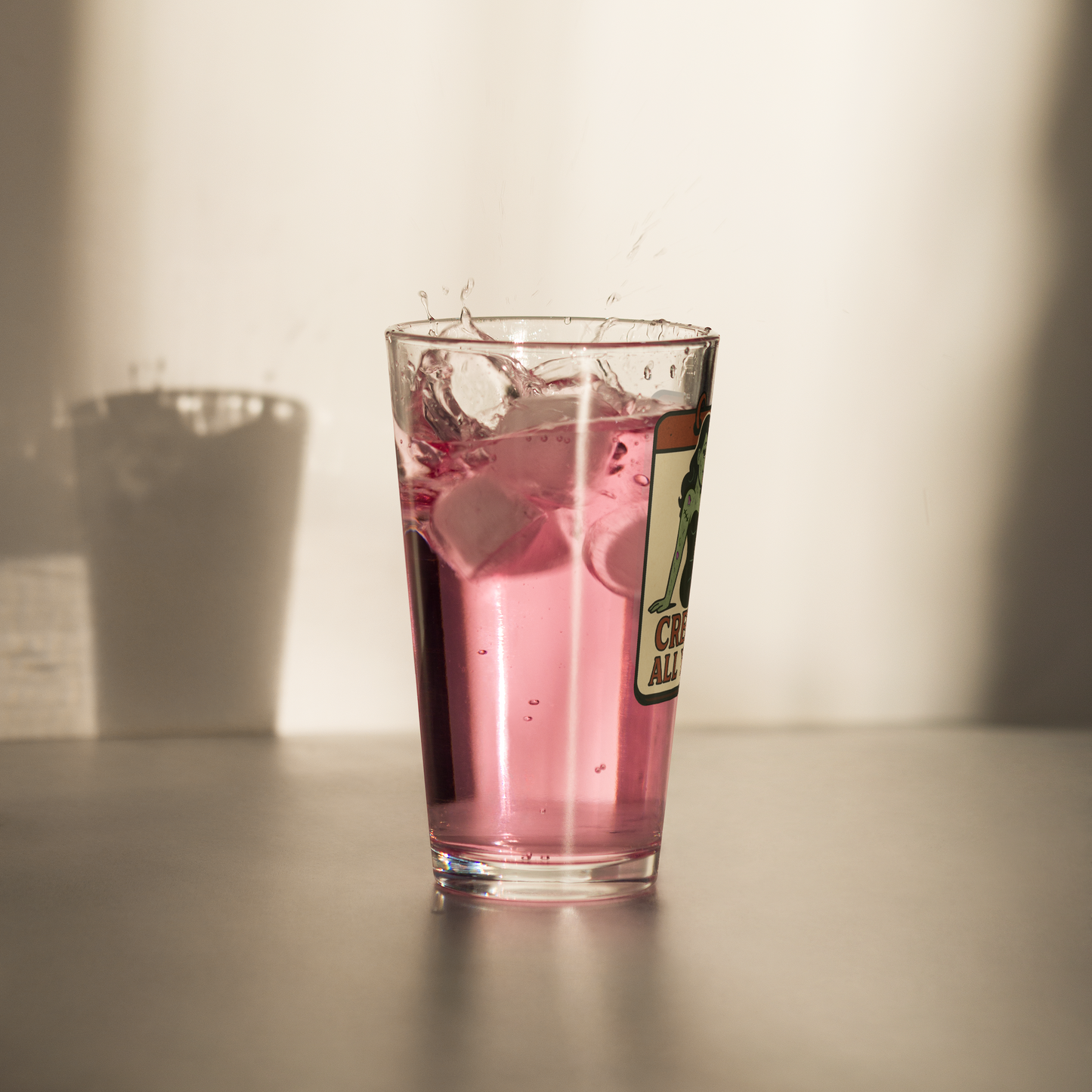 Glass of pink drink with ice cubes on a neutral background. Pint glass with a graphic design that features a green pinup-style zombie girl holding flowers, with a banner underneath that reads “CREEPY ALL YEAR.”