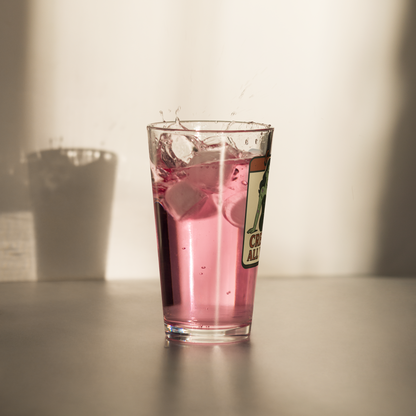 Glass of pink drink with ice cubes on a neutral background. Pint glass with a graphic design that features a green pinup-style zombie girl holding flowers, with a banner underneath that reads “CREEPY ALL YEAR.”
