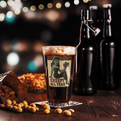 Pint of beer with a 'Creepy All Year' label on a wooden table with snacks and bottles in the background. The design features a green pinup-style zombie girl holding flowers, with a banner underneath that reads “CREEPY ALL YEAR.”
