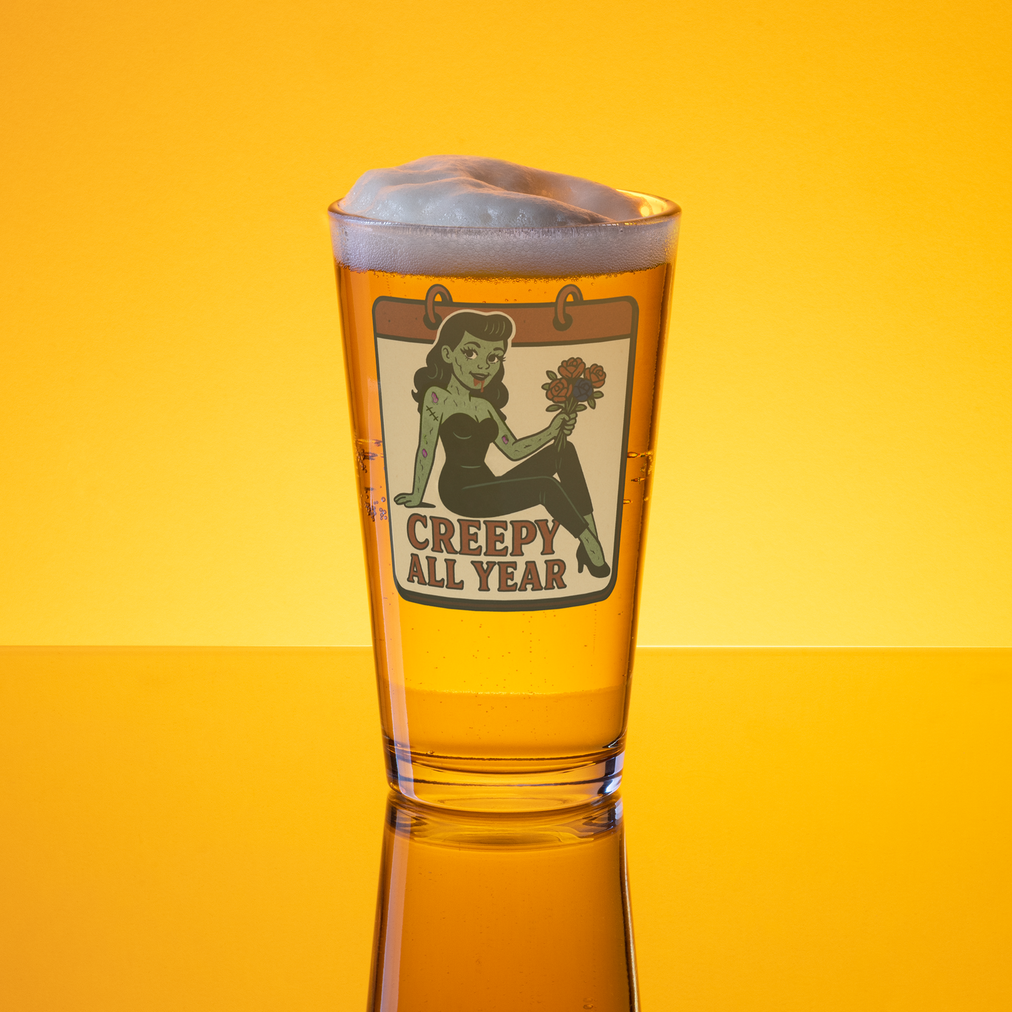 Beer glass with a 'Creepy All Year' label on a yellow background. The design features a green pinup-style zombie girl holding flowers, with a banner underneath that reads “CREEPY ALL YEAR.”