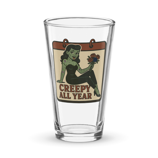 Pint glass with a graphic design. The design features a green pinup-style zombie girl holding flowers, with a banner underneath that reads “CREEPY ALL YEAR.”