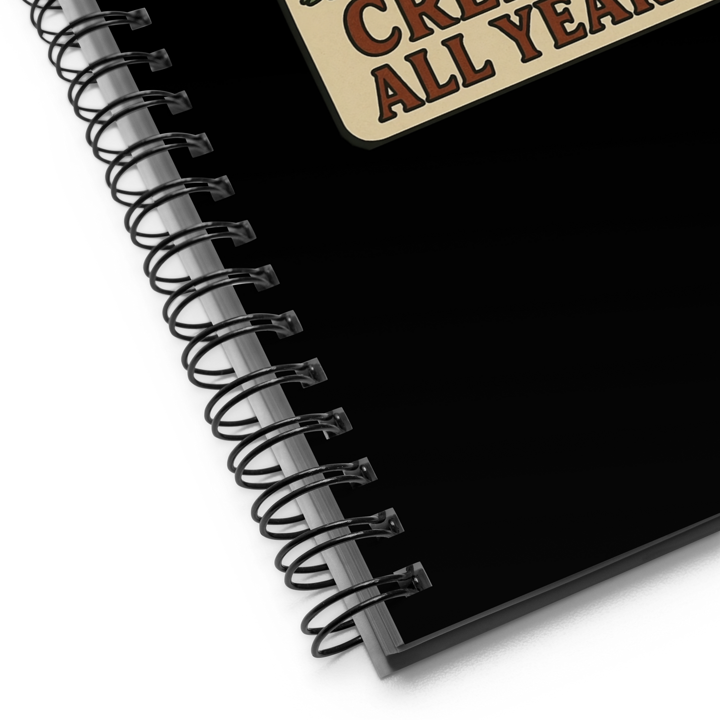 Black spiral-bound notebook with a decorative corner label on a white background. The design has a banner underneath that reads “CREEPY ALL YEAR.”
