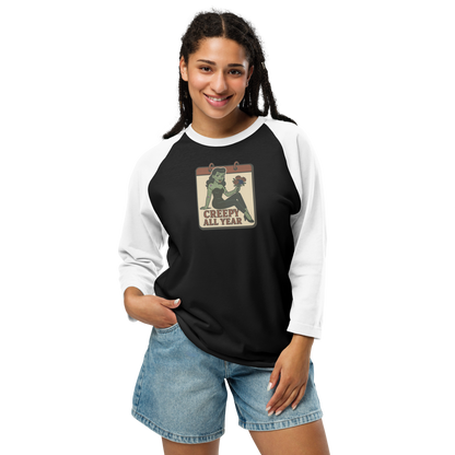 A woman with black hair wearing jean shorts and black and white raglan shirt. The design features a green pinup-style zombie girl holding flowers, with a banner underneath that reads “CREEPY ALL YEAR.”