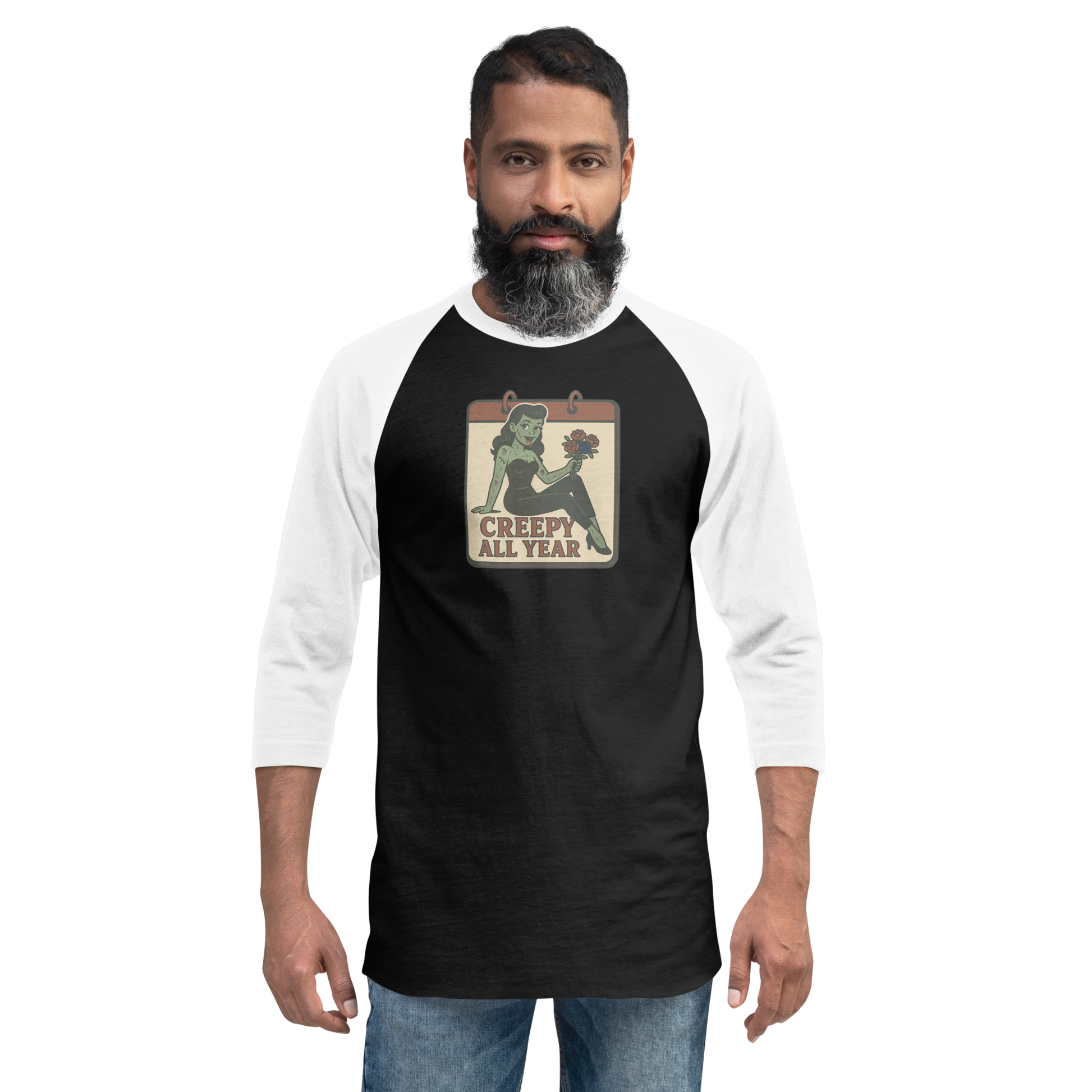 A man with black hair wearing jeans and a black and white raglan t-shirt with a small vintage-style design on the pocket area. The design features a green pinup-style zombie girl holding flowers, with a banner underneath that reads “CREEPY ALL YEAR.”