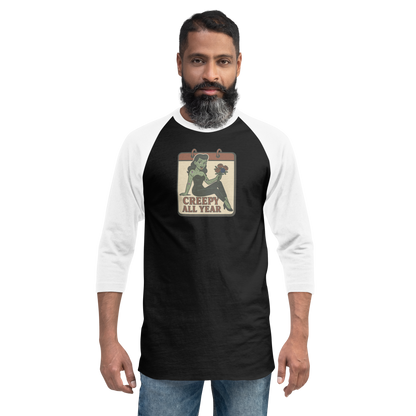 A man with black hair wearing jeans and a black and white raglan t-shirt with a small vintage-style design on the pocket area. The design features a green pinup-style zombie girl holding flowers, with a banner underneath that reads “CREEPY ALL YEAR.”