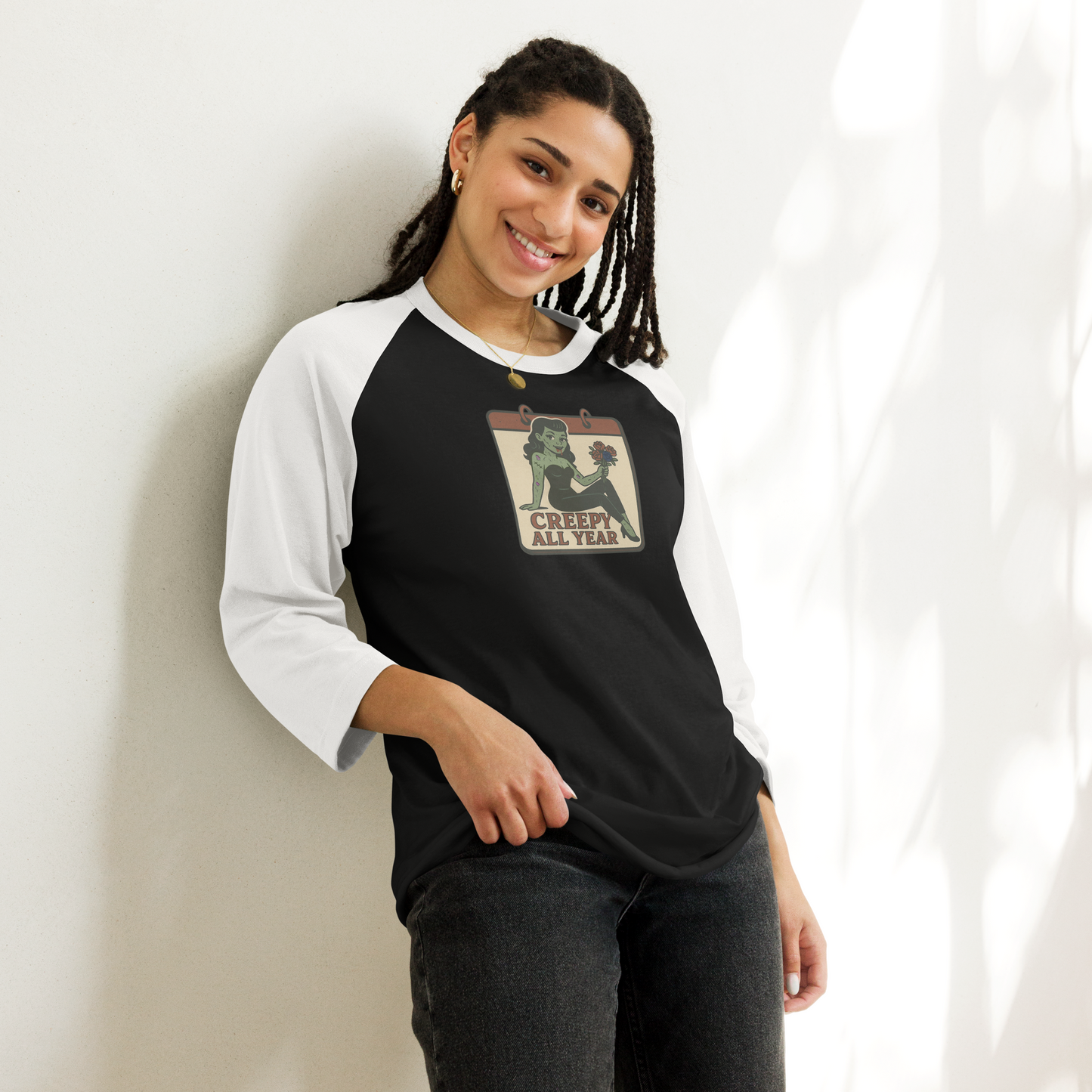 A woman with black hair wearing dark jeans and a black and white raglan t-shirt with a vintage-style design on the pocket area. The design features a green pinup-style zombie girl holding flowers, with a banner underneath that reads “CREEPY ALL YEAR.”
