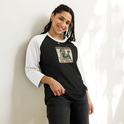 A woman with black hair wearing dark jeans and a black and white raglan t-shirt with a vintage-style design on the pocket area. The design features a green pinup-style zombie girl holding flowers, with a banner underneath that reads “CREEPY ALL YEAR.”
