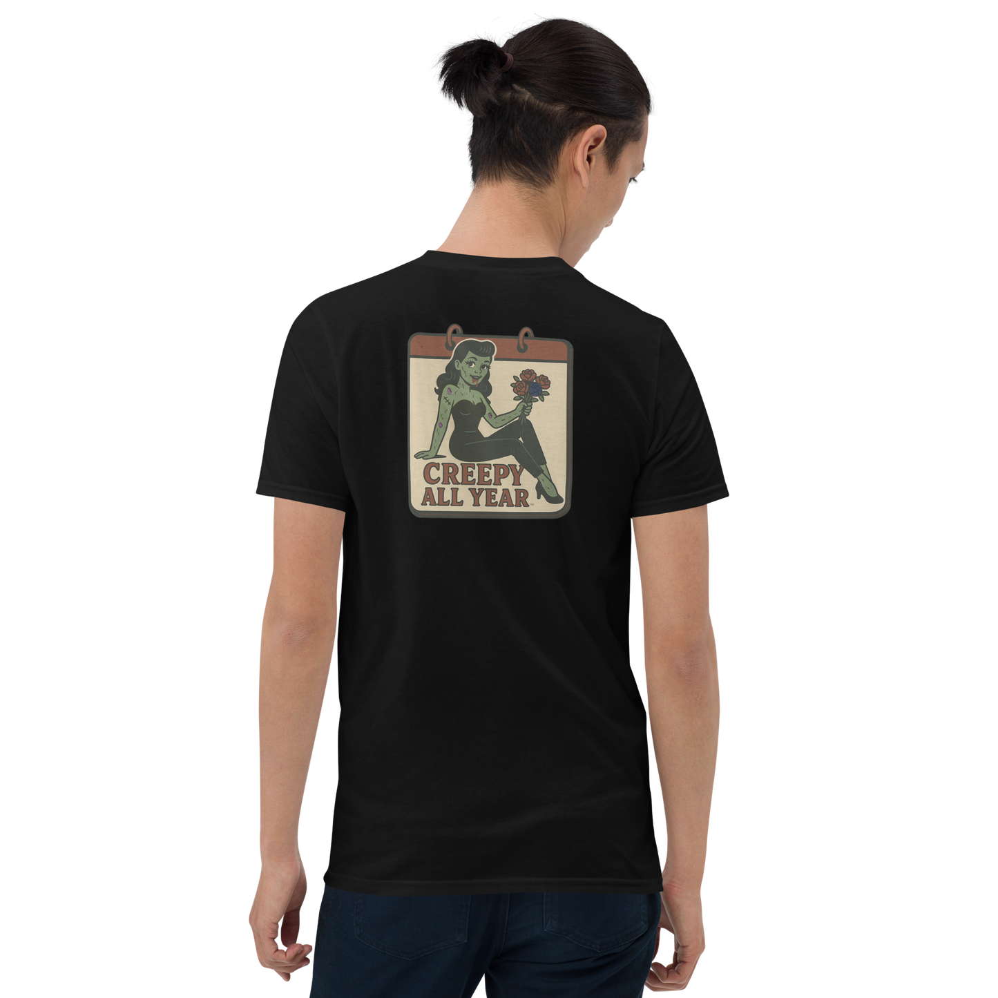 A black t-shirt with a vintage-style design on the back area work by a man with dark hair in a ponytail. The design features a green pinup-style zombie girl holding flowers, with a banner underneath that reads “CREEPY ALL YEAR.”