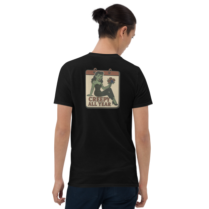 A black t-shirt with a vintage-style design on the back area work by a man with dark hair in a ponytail. The design features a green pinup-style zombie girl holding flowers, with a banner underneath that reads “CREEPY ALL YEAR.”