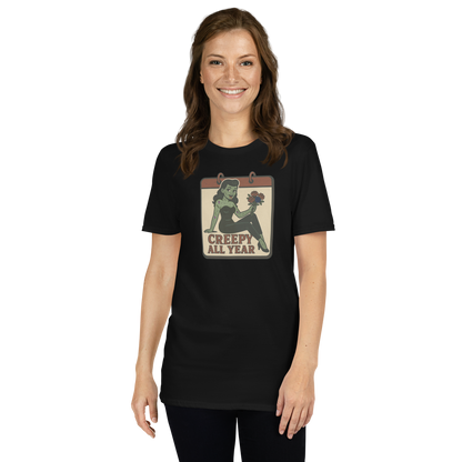 A t-shirt on a girl with brown hair with a vintage-style design on the front. The design features a green pinup-style zombie girl holding flowers, with a banner underneath that reads “CREEPY ALL YEAR.”