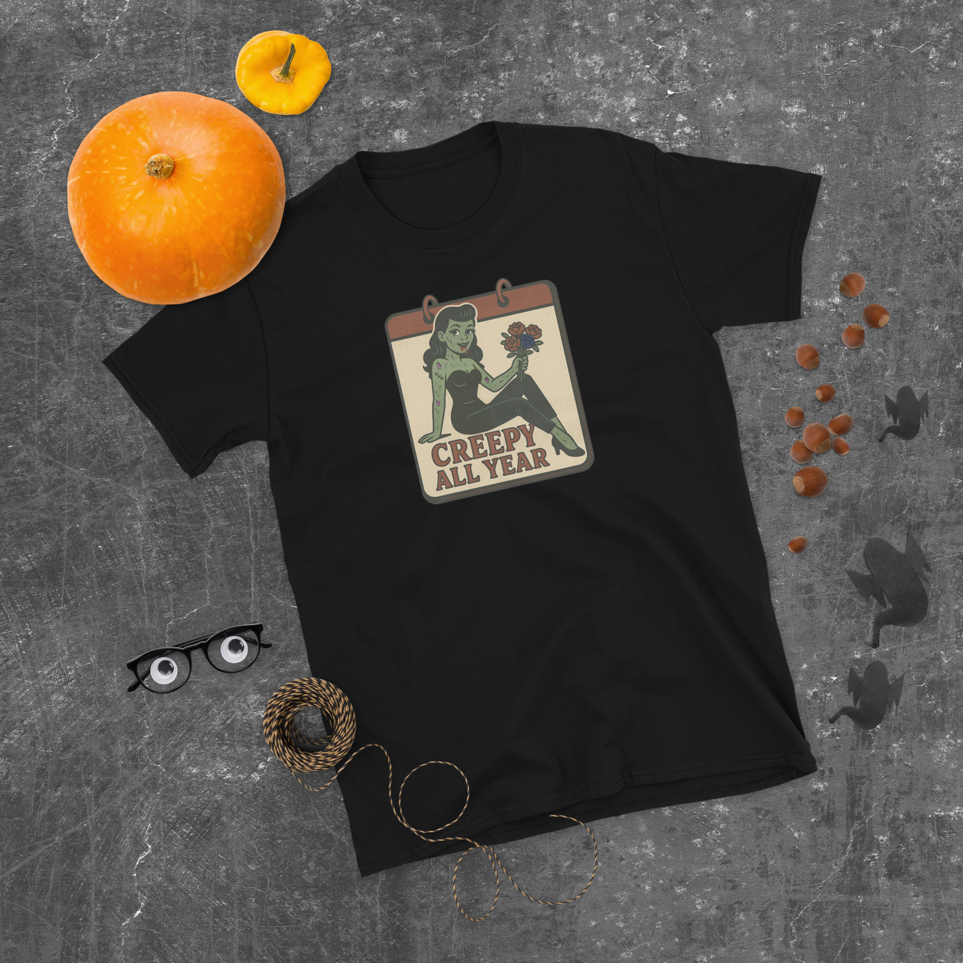 Black t-shirt with Halloween-themed graphic on a gray surface with pumpkins and decorative items. The design features a green pinup-style zombie girl holding flowers, with a banner underneath that reads “CREEPY ALL YEAR.”