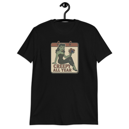 A black t-shirt on a hanger with a vintage-style design on the front. The design features a green pinup-style zombie girl holding flowers, with a banner underneath that reads “CREEPY ALL YEAR.”