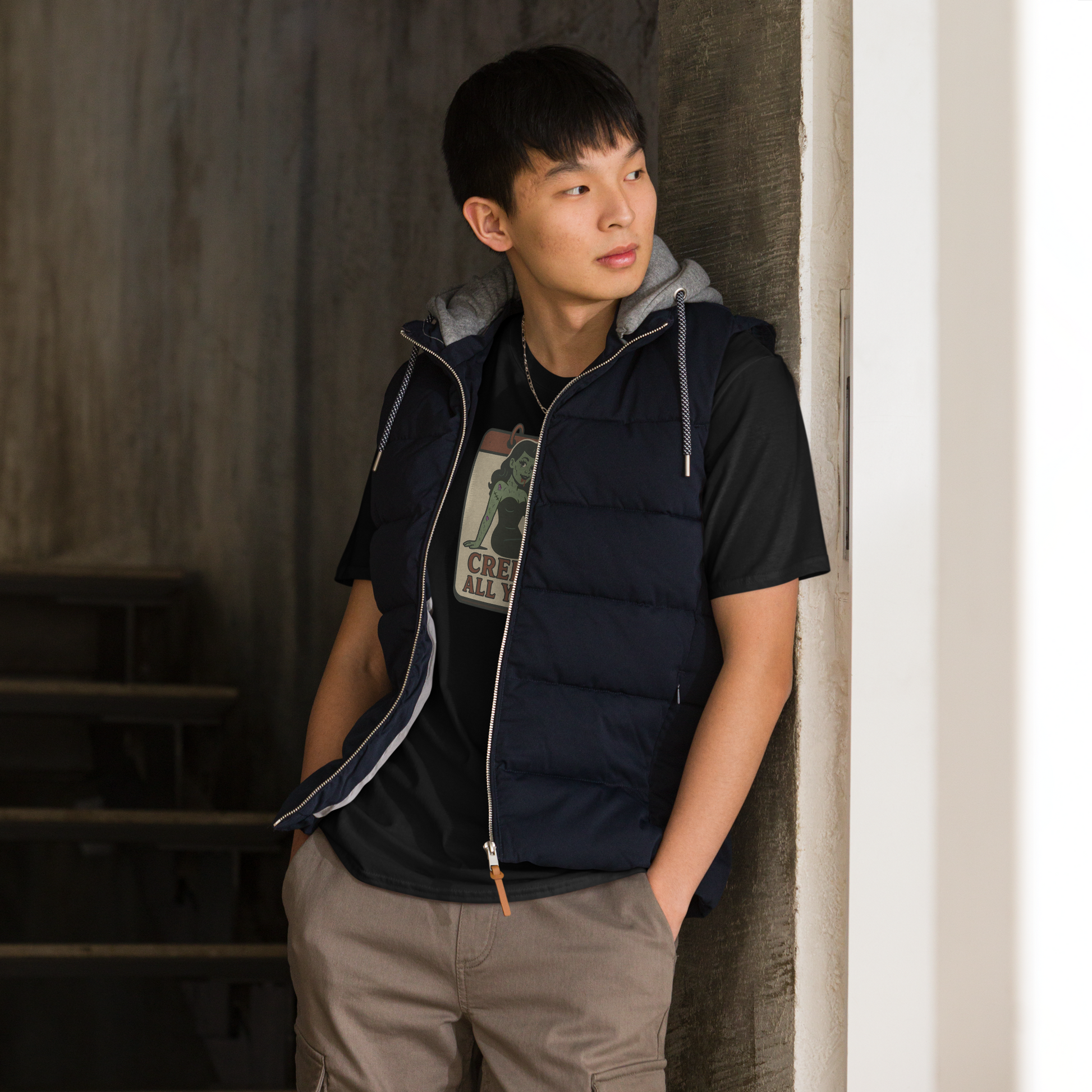 A man indoors standing in front of stairs with black hair wearing cargo pants and t-shirt with vintage style design. The design on the front features a green pinup-style zombie girl holding flowers, with a banner underneath that reads “CREEPY ALL YEAR.”