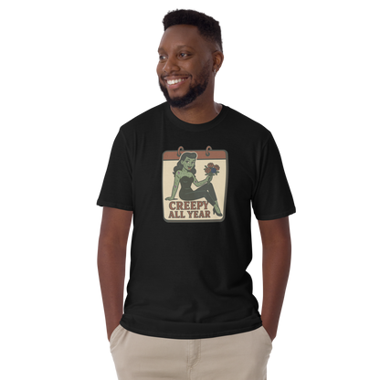 A t-shirt on a man with short balck hair with a vintage-style design on the front. The design features a green pinup-style zombie girl holding flowers, with a banner underneath that reads “CREEPY ALL YEAR.”