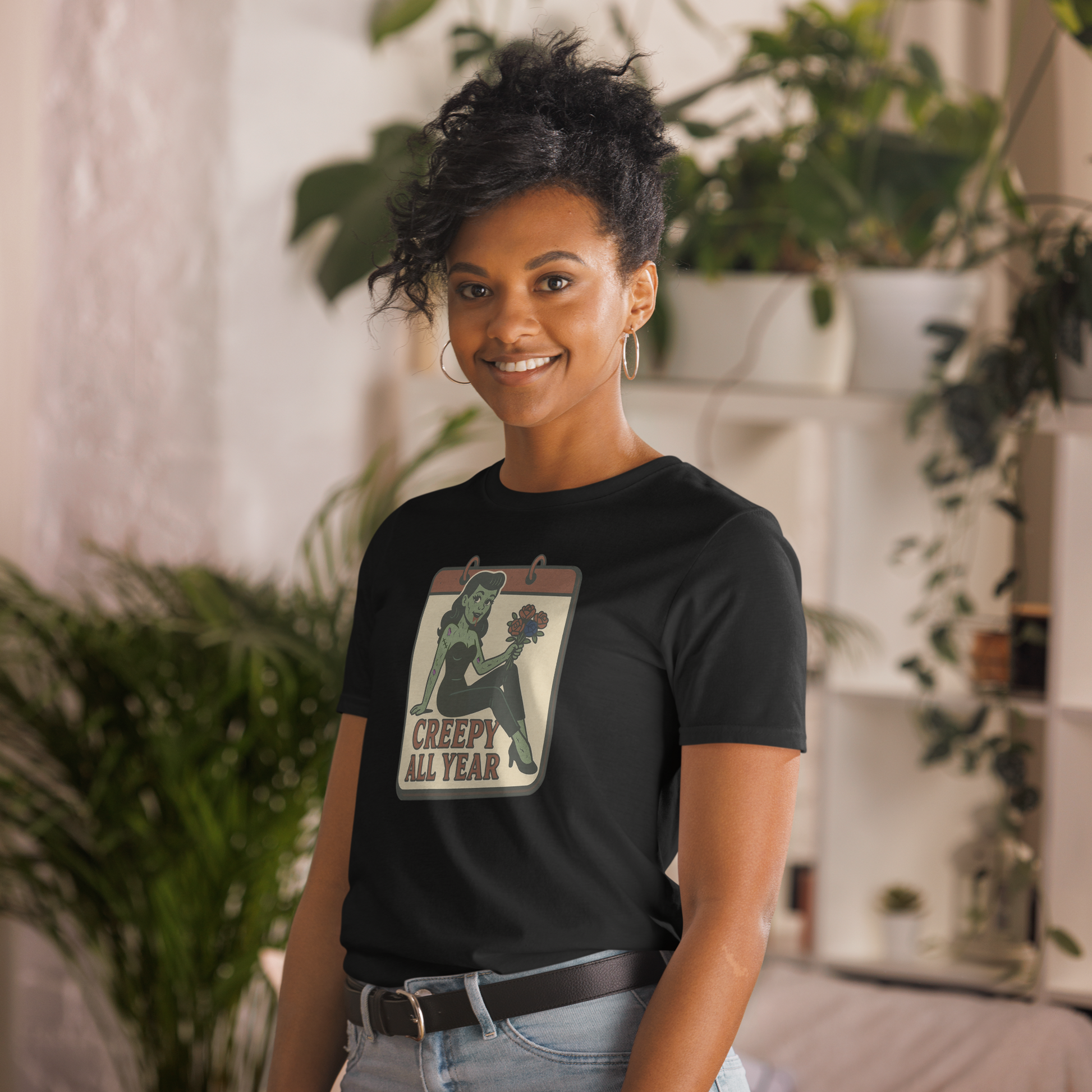 A girl indoors surrounded by plants with black hair wearing jeans and t-shirt with vintage style design. The design on the front features a green pinup-style zombie girl holding flowers, with a banner underneath that reads “CREEPY ALL YEAR.”