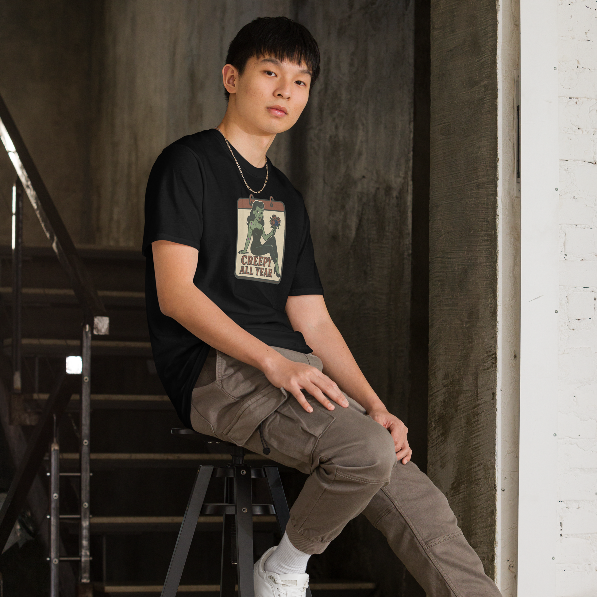 A man indoors sitting on a black stool with black hair wearing cargo pants and t-shirt with vintage style design. The design on the front features a green pinup-style zombie girl holding flowers, with a banner underneath that reads “CREEPY ALL YEAR.”