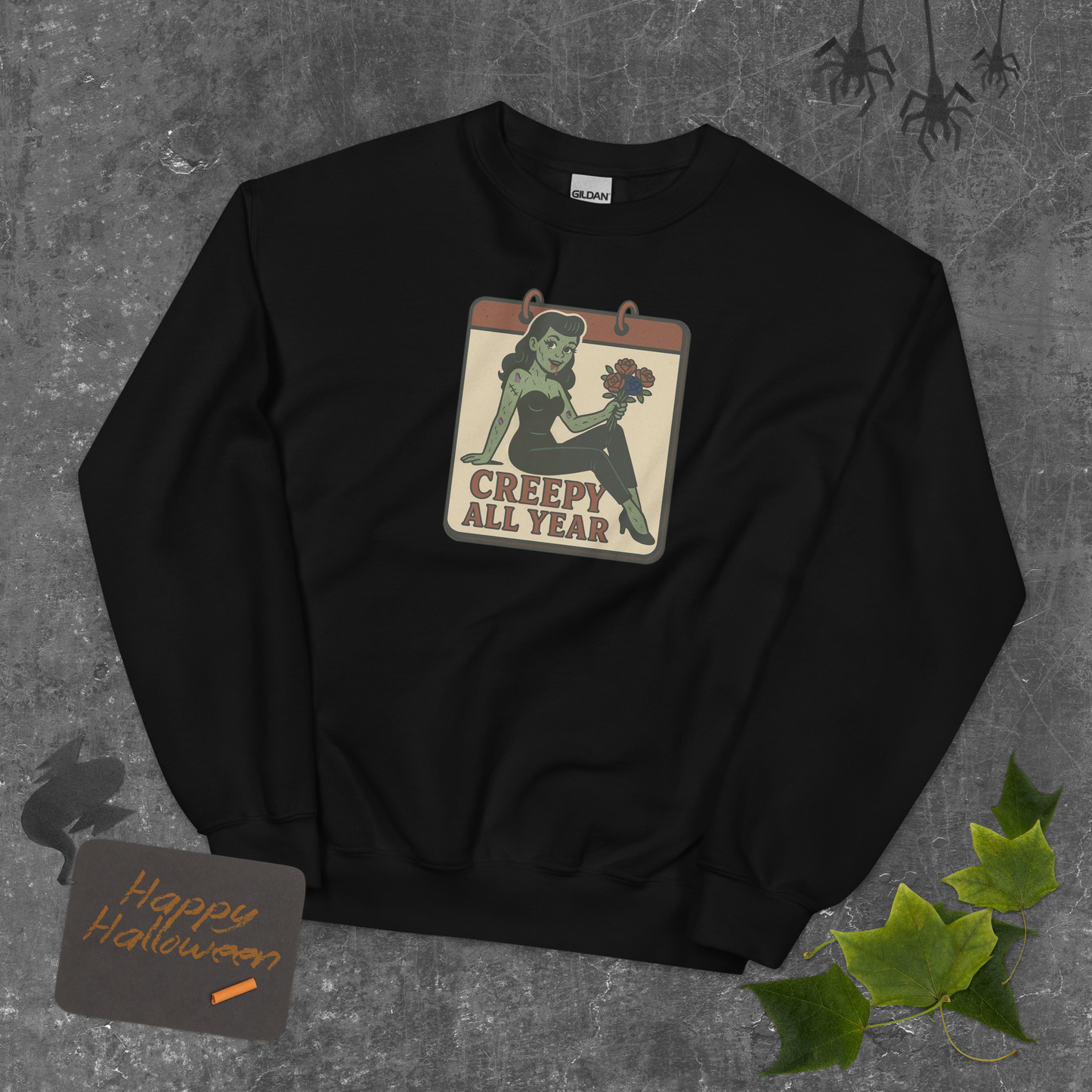 Black sweatshirt with a graphic design on a textured gray background. The design features a green pinup-style zombie girl holding flowers, with a banner underneath that reads “CREEPY ALL YEAR.”