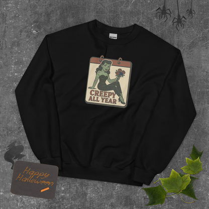Black sweatshirt with a graphic design on a textured gray background. The design features a green pinup-style zombie girl holding flowers, with a banner underneath that reads “CREEPY ALL YEAR.”