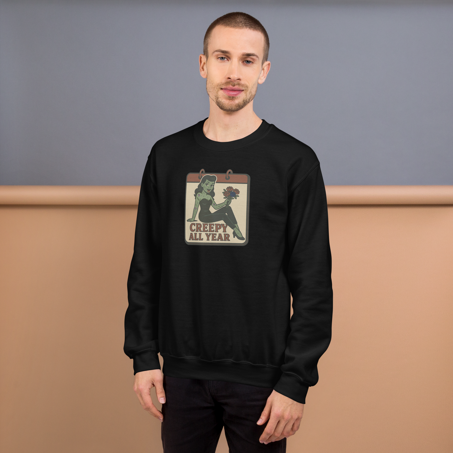 A black sweatshirt on a man with short brown hair with a vintage-style design on the front. The design features a green pinup-style zombie girl holding flowers, with a banner underneath that reads “CREEPY ALL YEAR.”