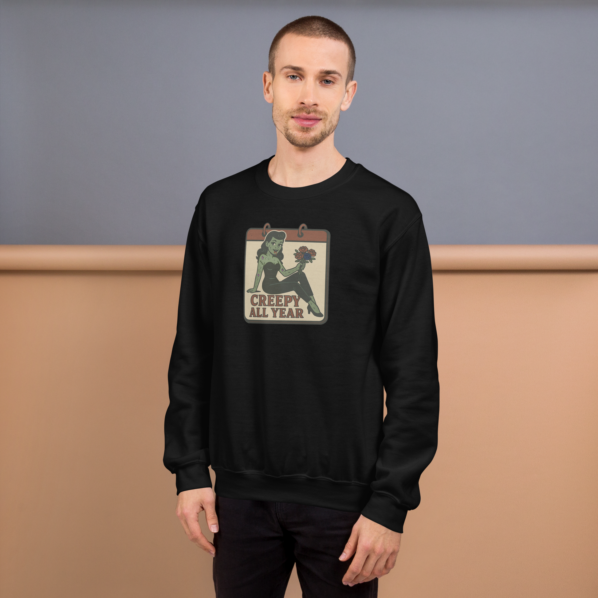 A black sweatshirt on a man with short brown hair with a vintage-style design on the front. The design features a green pinup-style zombie girl holding flowers, with a banner underneath that reads “CREEPY ALL YEAR.”