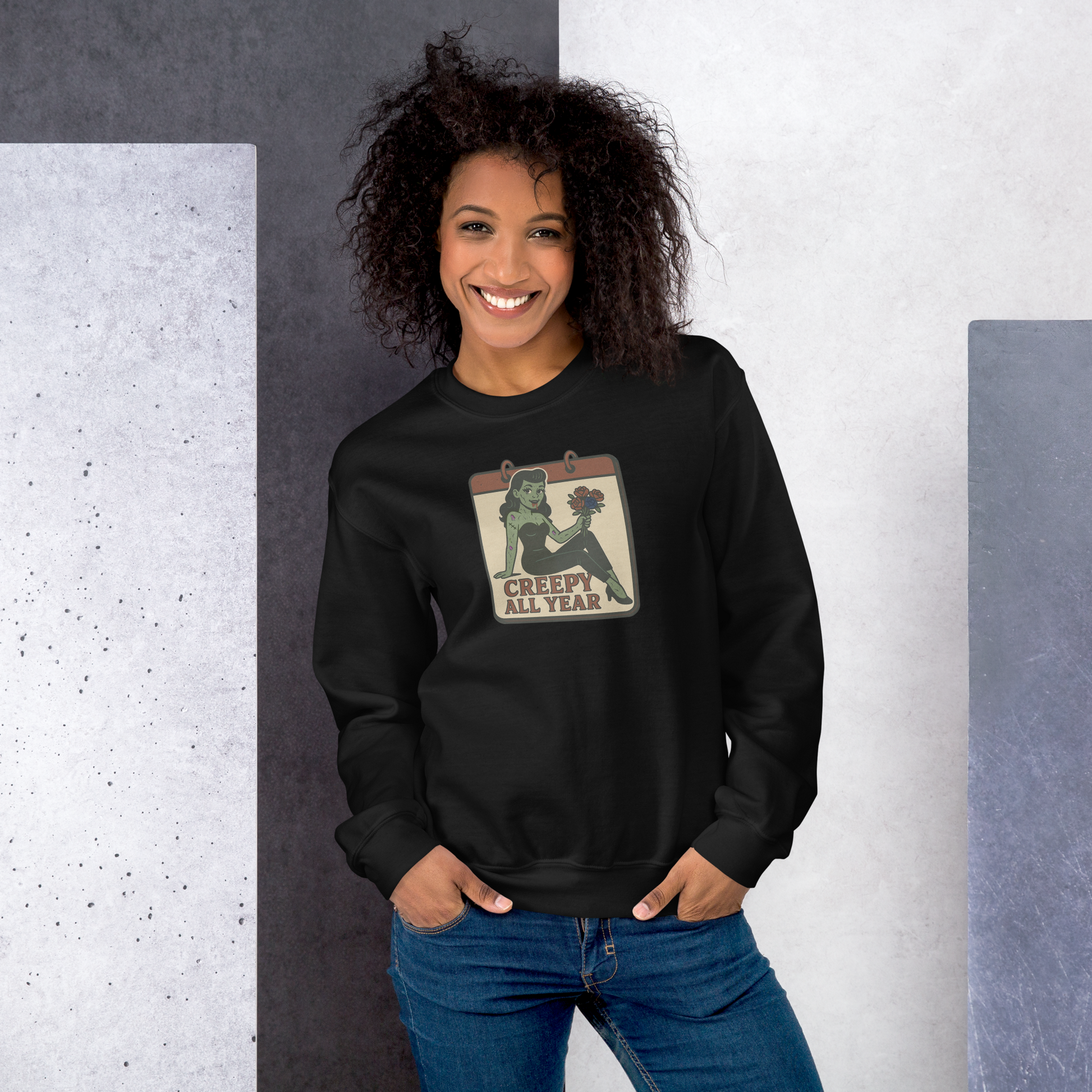 A black sweatshirt on a girl with black hair with a vintage-style design on the front. The design features a green pinup-style zombie girl holding flowers, with a banner underneath that reads “CREEPY ALL YEAR.”