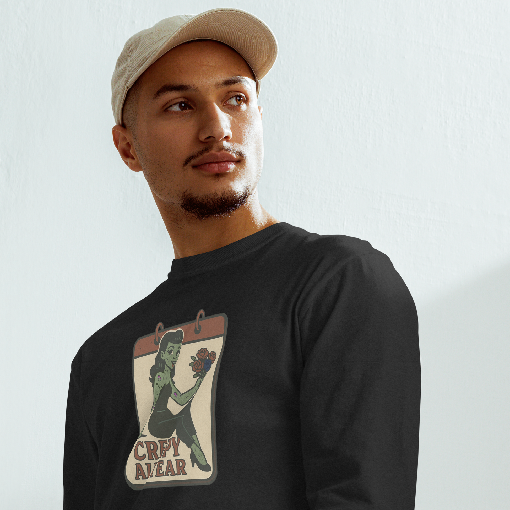 A man with a light baseball hat wearing a black long sleeve t-shirt. The design on the front features a green pinup-style zombie girl holding flowers, with a banner underneath that reads “CREEPY ALL YEAR.”