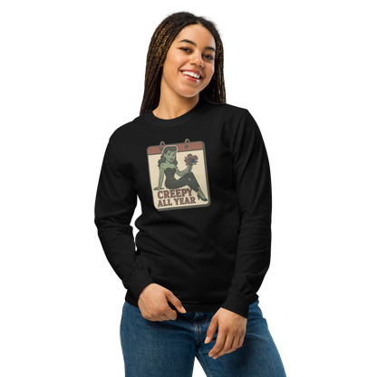 A girl smiling with braids wearing jeans and a black long sleeve t-shirt. The design on the front features a green pinup-style zombie girl holding flowers, with a banner underneath that reads “CREEPY ALL YEAR.”