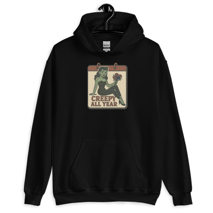 A black hoodie on a hanger with a vintage-style design on the front. The design features a green pinup-style zombie girl holding flowers, with a banner underneath that reads “CREEPY ALL YEAR.”