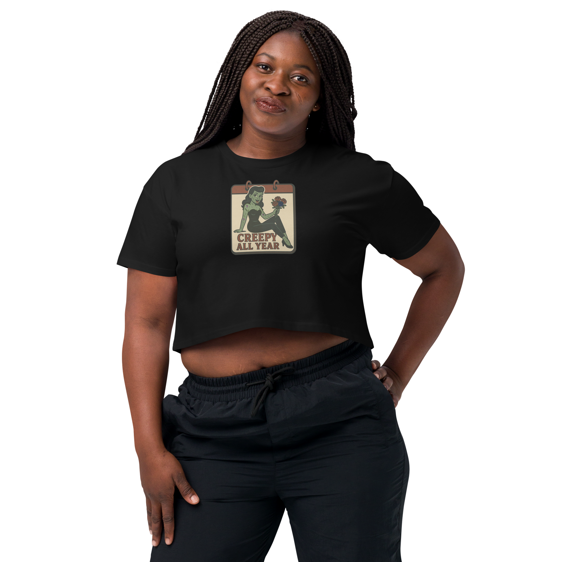 A black crop top on a woman with braids, with a vintage-style design on the front. The design features a green pinup-style zombie girl holding flowers, with a banner underneath that reads “CREEPY ALL YEAR.”