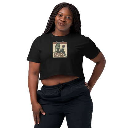A black crop top on a woman with braids, with a vintage-style design on the front. The design features a green pinup-style zombie girl holding flowers, with a banner underneath that reads “CREEPY ALL YEAR.”