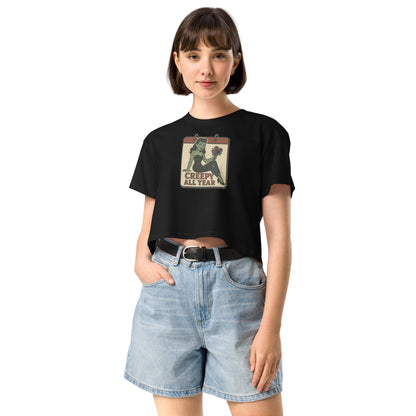 A black crop top on a girl with dark hair and jean shorts with a vintage-style design on the front. The design features a green pinup-style zombie girl holding flowers, with a banner underneath that reads “CREEPY ALL YEAR.”