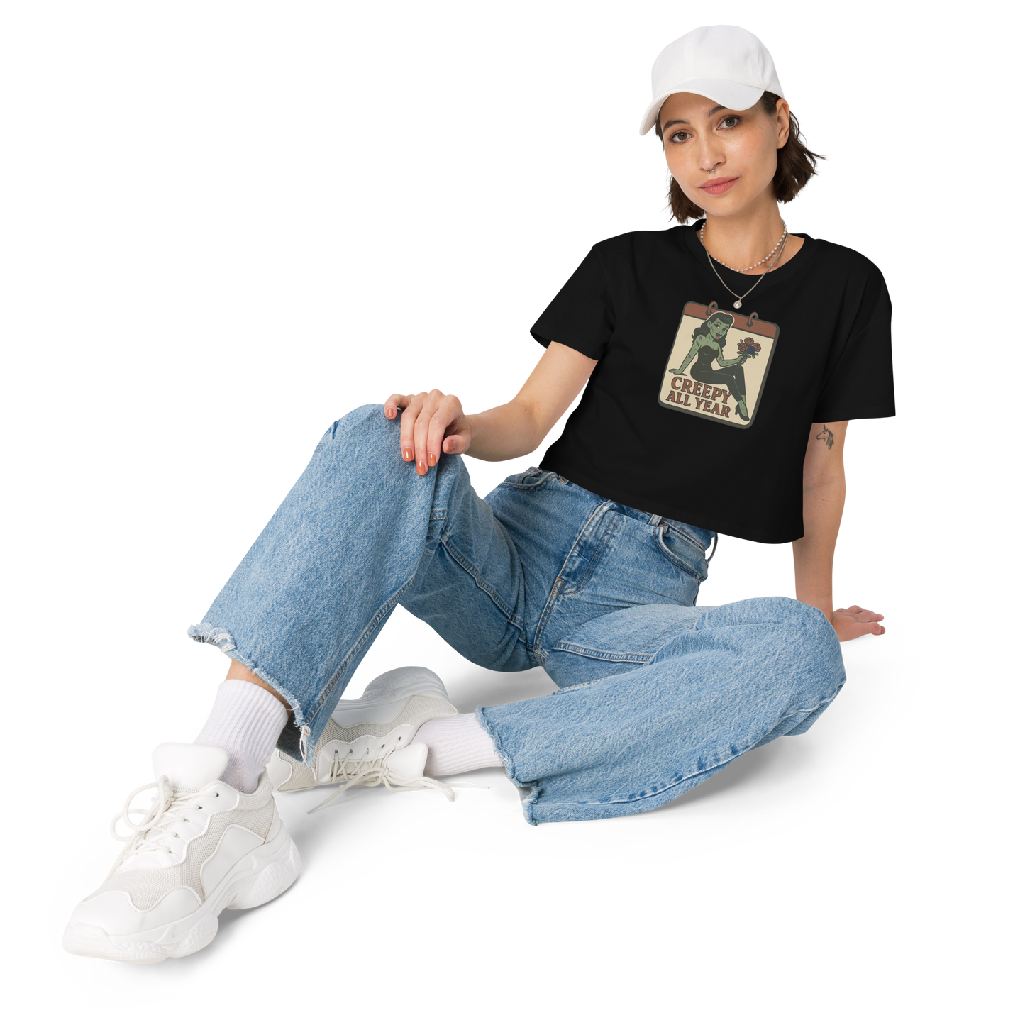 A black crop top on a girl with dark hair, jeans, white hat with a vintage-style design on the front. The design features a green pinup-style zombie girl holding flowers, with a banner underneath that reads “CREEPY ALL YEAR.”