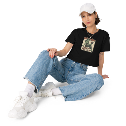 A black crop top on a girl with dark hair, jeans, white hat with a vintage-style design on the front. The design features a green pinup-style zombie girl holding flowers, with a banner underneath that reads “CREEPY ALL YEAR.”