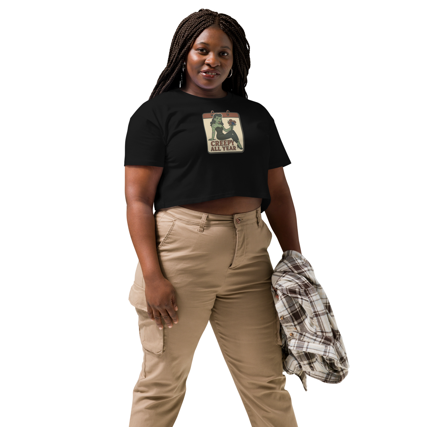 A black crop top on a woman with braids, tan pants, with a vintage-style design on the front. The design features a green pinup-style zombie girl holding flowers, with a banner underneath that reads “CREEPY ALL YEAR.”