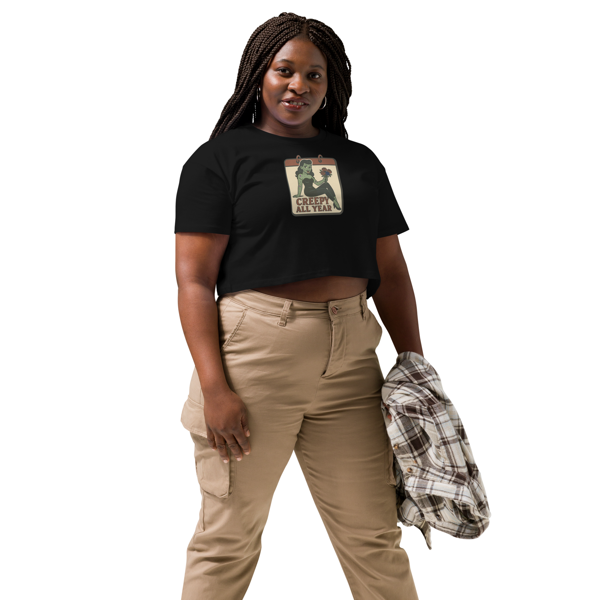 A black crop top on a woman with braids, tan pants, with a vintage-style design on the front. The design features a green pinup-style zombie girl holding flowers, with a banner underneath that reads “CREEPY ALL YEAR.”