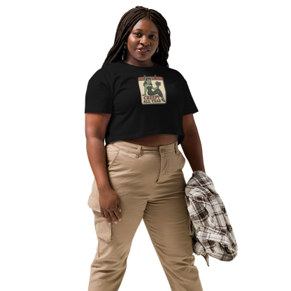 A black crop top on a woman with braids, tan pants, with a vintage-style design on the front. The design features a green pinup-style zombie girl holding flowers, with a banner underneath that reads “CREEPY ALL YEAR.”