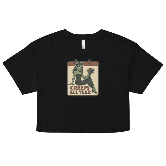 A black crop top with a vintage-style design on the front. The design features a green pinup-style zombie girl holding flowers, with a banner underneath that reads “CREEPY ALL YEAR.”