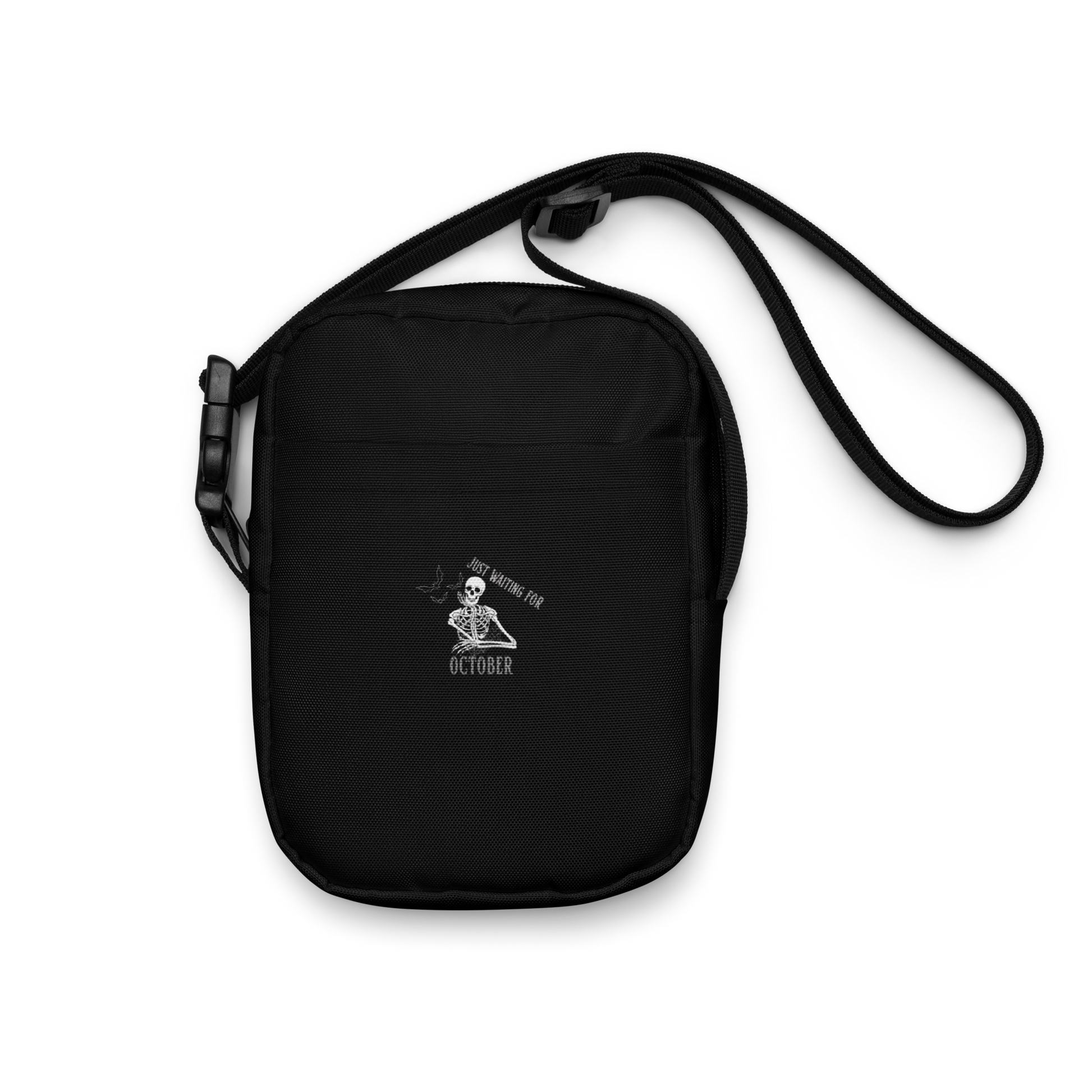 This is a black crossbody bag that has a bored skeleton with his head resting in his hand and he has spider webs and bats around him and the text reads just waiting for October