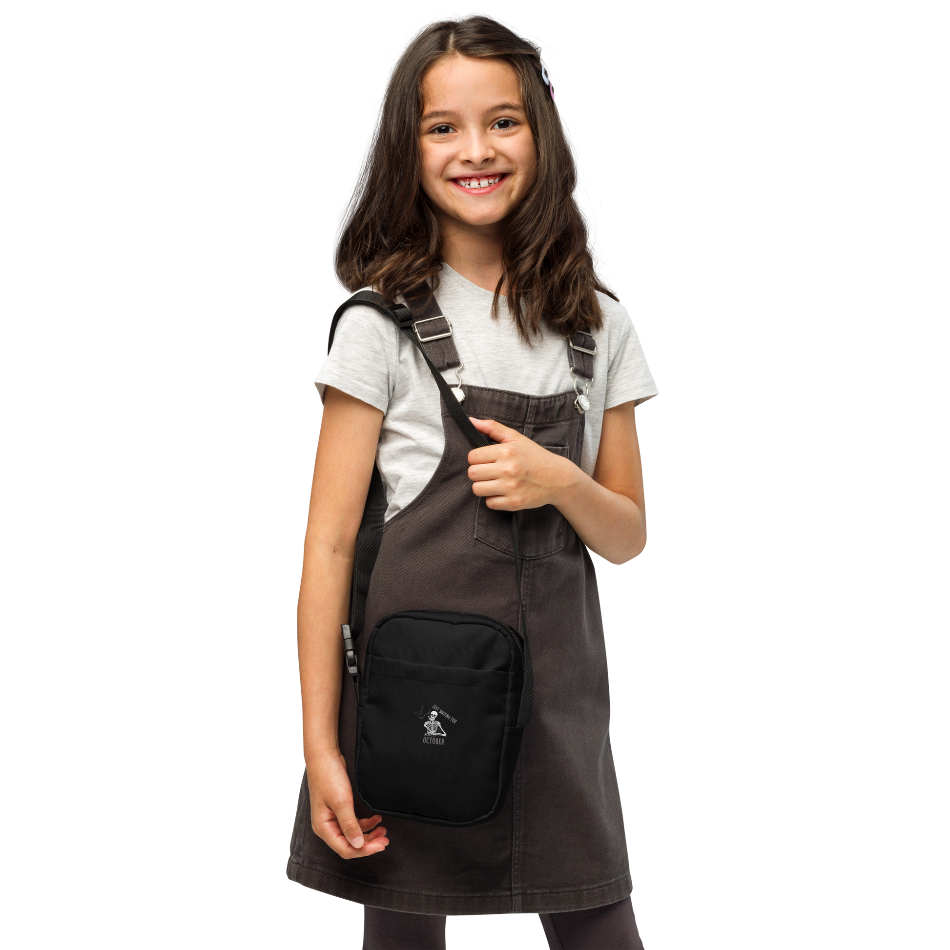 This is a kid with dark hair wearing a black crossbody bag that has a bored skeleton with his head resting in his hand and he has spider webs and bats around him and the text reads just waiting for October