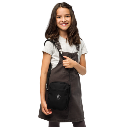 This is a kid with dark hair wearing a black crossbody bag that has a bored skeleton with his head resting in his hand and he has spider webs and bats around him and the text reads just waiting for October