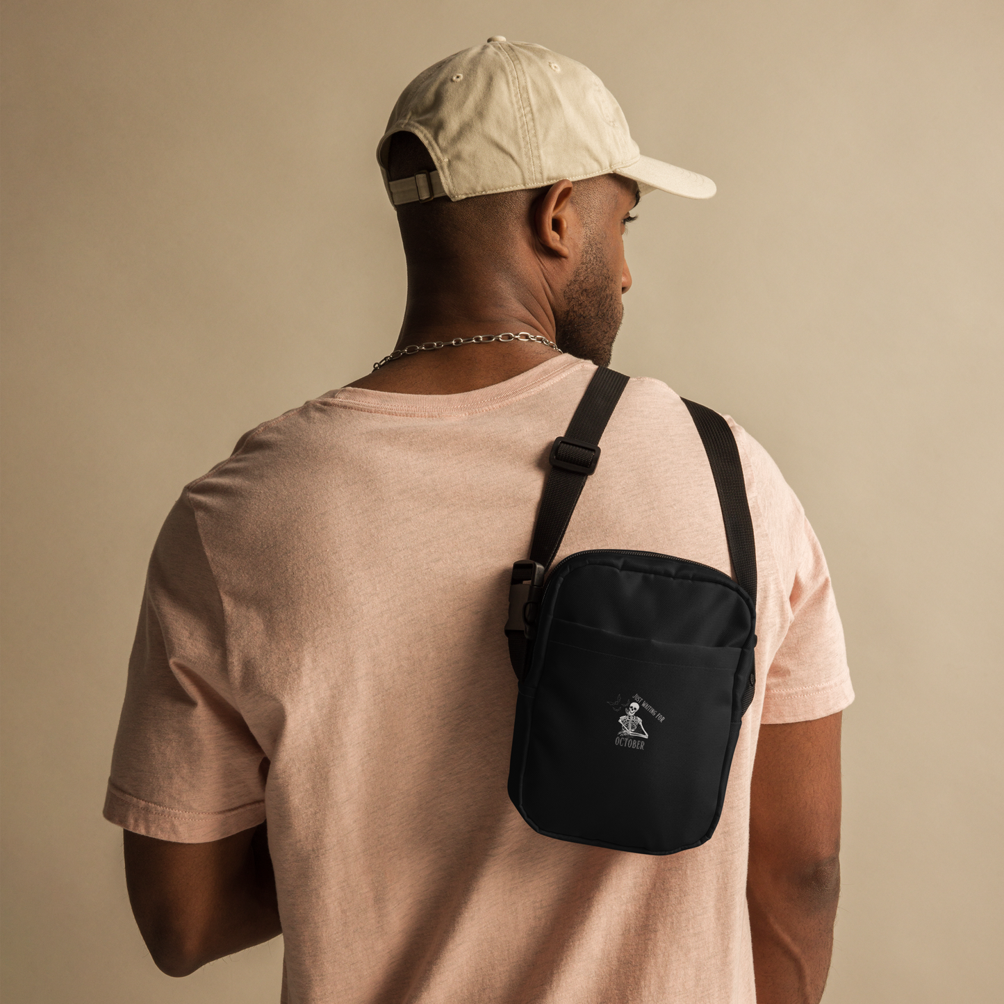 This is a man with a baseball hat holding a black crossbody bag that has a bored skeleton with his head resting in his hand and he has spider webs and bats around him and the text reads just waiting for October