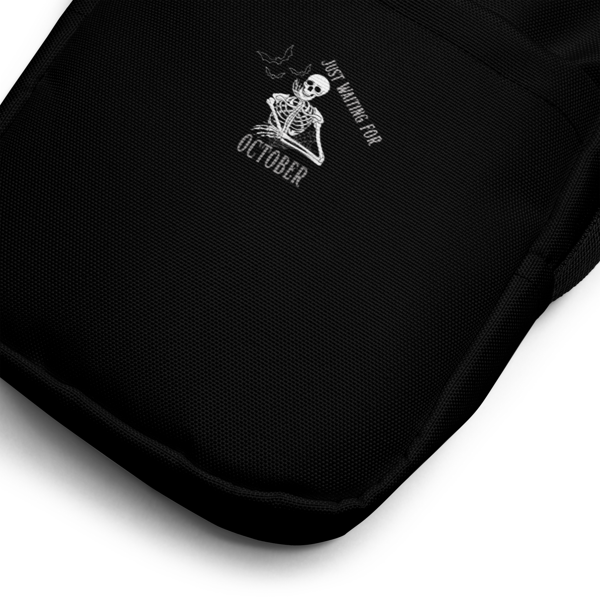 This is a zoom in of a black crossbody bag that has a bored skeleton with his head resting in his hand and he has spider webs and bats around him and the text reads just waiting for October