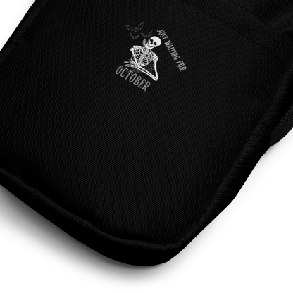 This is a zoom in of a black crossbody bag that has a bored skeleton with his head resting in his hand and he has spider webs and bats around him and the text reads just waiting for October